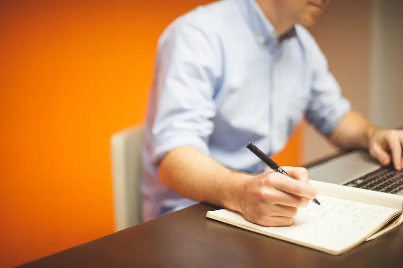 Homem sentado em uma cadeira de frente para mesa escrevendo em um caderno observações da numerologia empresarial