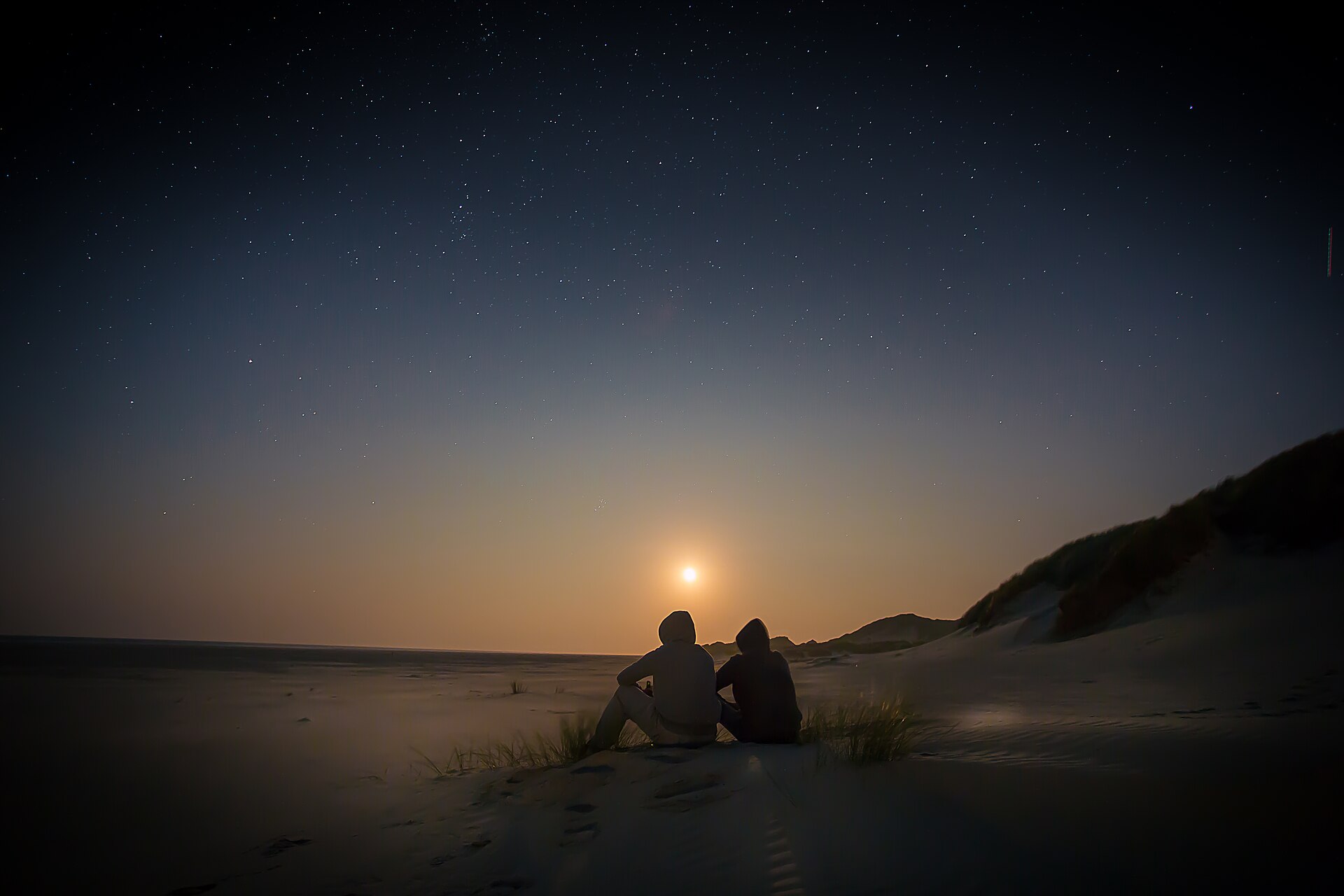 Duas pessoas sentadas de costas numa duna, olhando o poente sob um céu estrelado